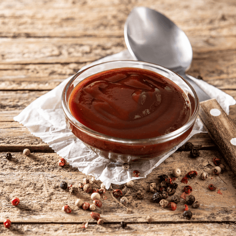 barbecue sauce in glass dish next to metal spoon surrounded by peppercorns